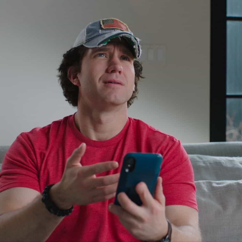 A young man in a red T-shirt and cap talks while holding his phone, from a Selective Service PSA