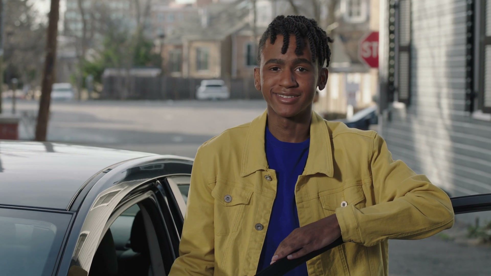 A young man in a yellow jacket leans on his car outside a row of brick houses and smiles at camera.