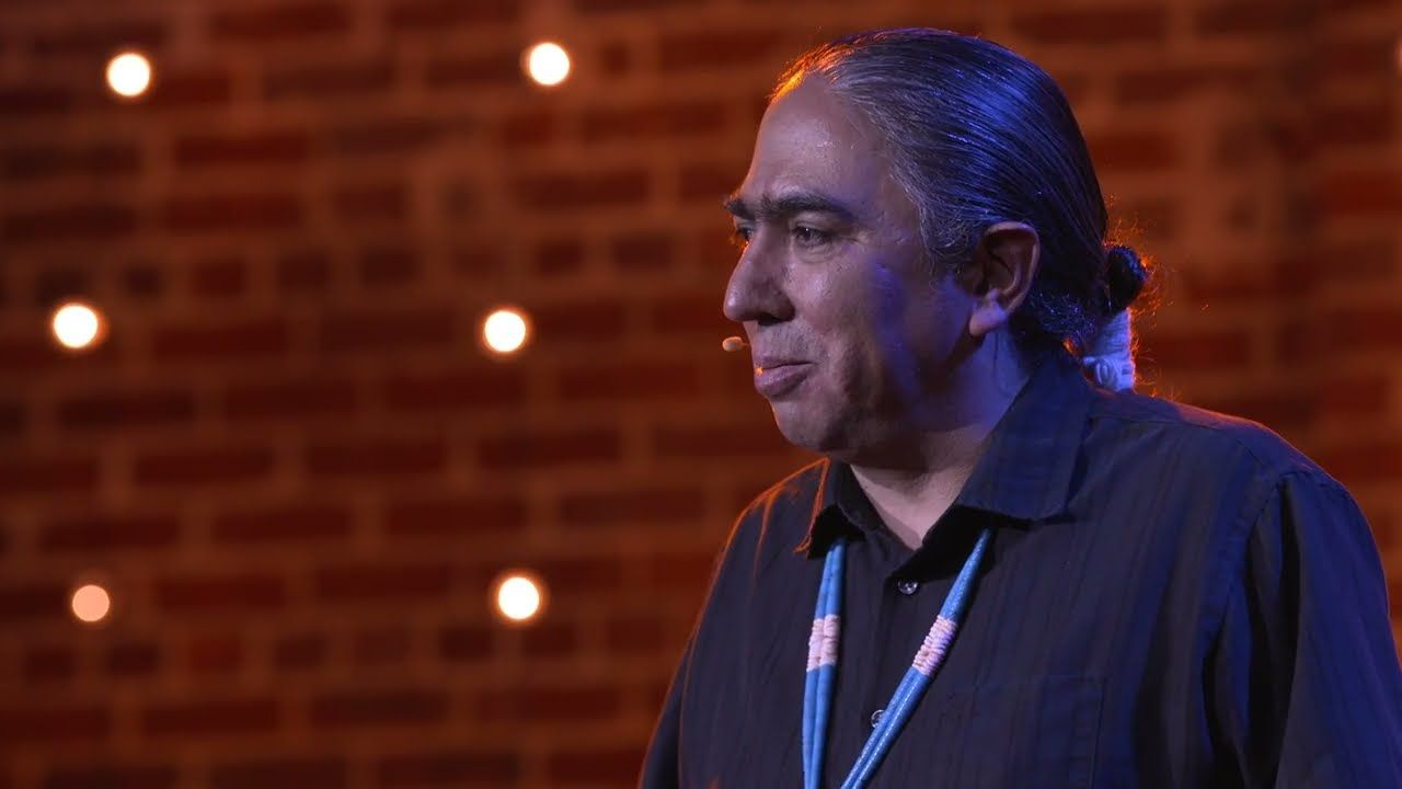 Speaker Mark Charles, wearing a headset mic and a lanyard, talks on stage in front of a brick wall with warm lights.