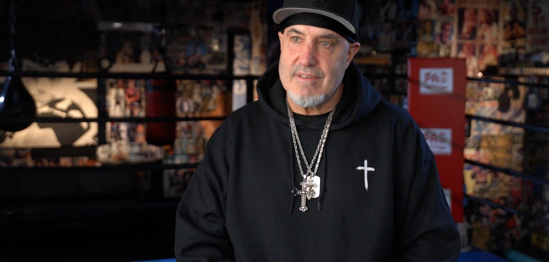 A man in a black cap and hoodie talks during an interview in a boxing gym.