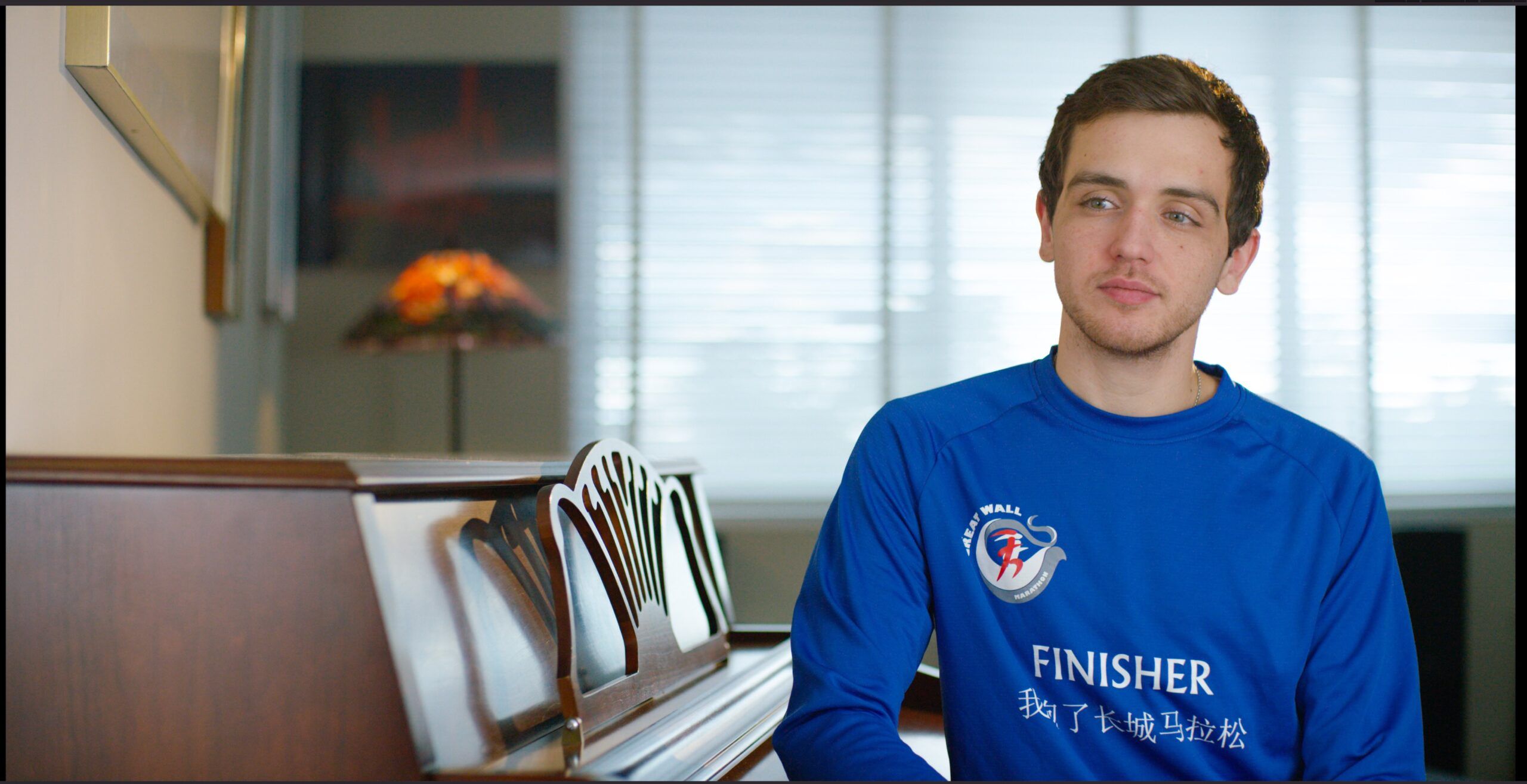 A young man in a blue marathon finisher shirt sits at a piano for an interview