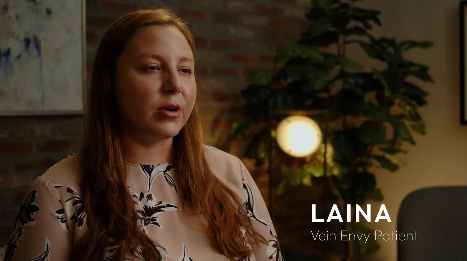 Laina, a Vein Envy patient with long red hair, tells her story on camera in front of a brick wall and a plant.