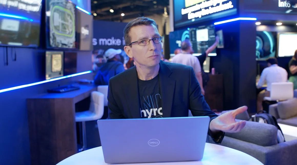 A presenter in glasses talks beside a laptop in a trade show booth lit in blue
