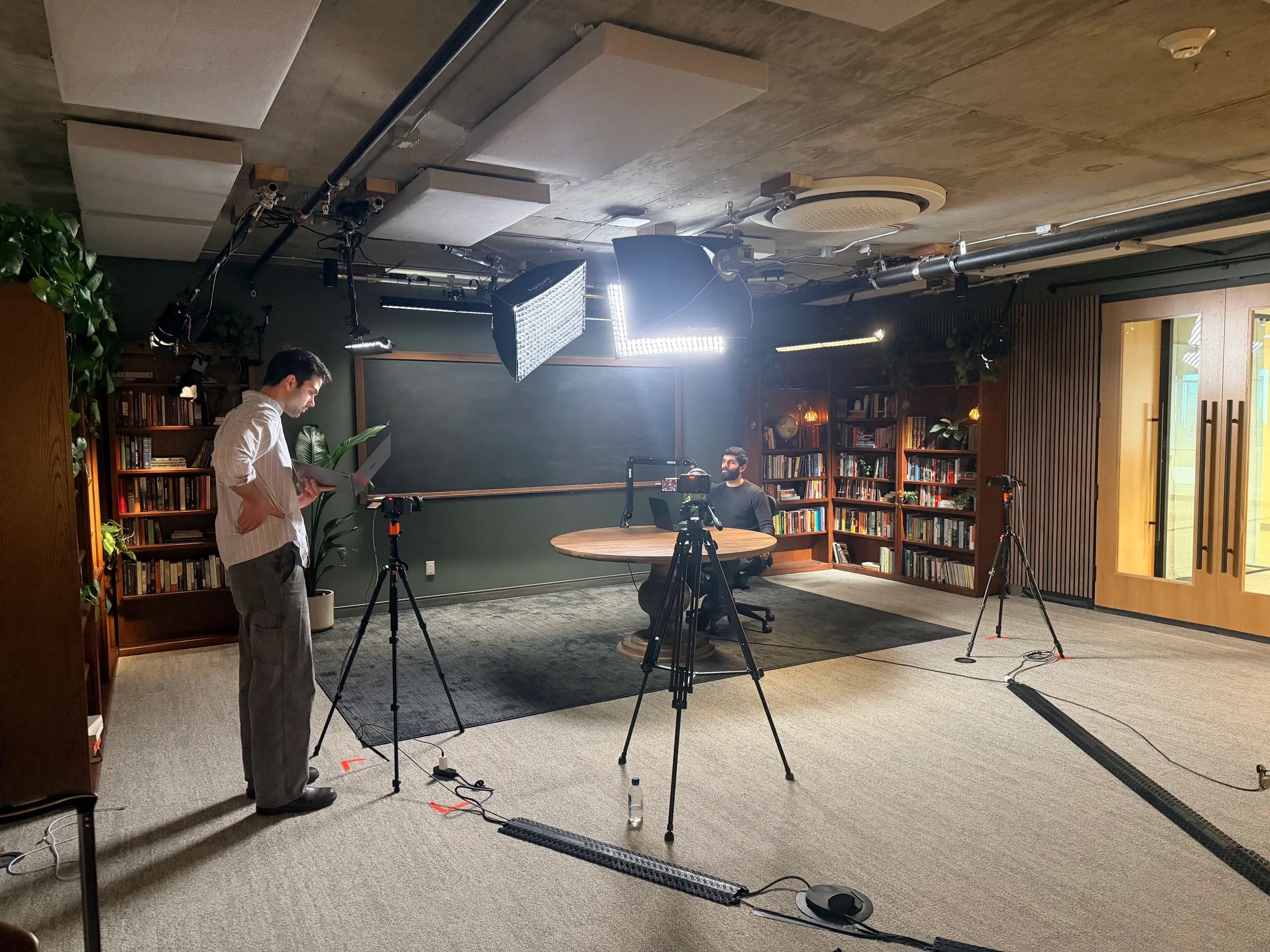 A crew member checks a tablet beside cameras and a light on stands in a library studio