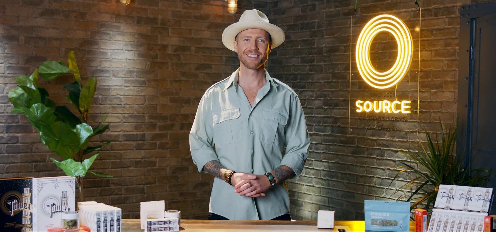 A presenter in a hat and pale shirt stands behind a product counter in a store, a neon Source sign on the brick wall behind him.