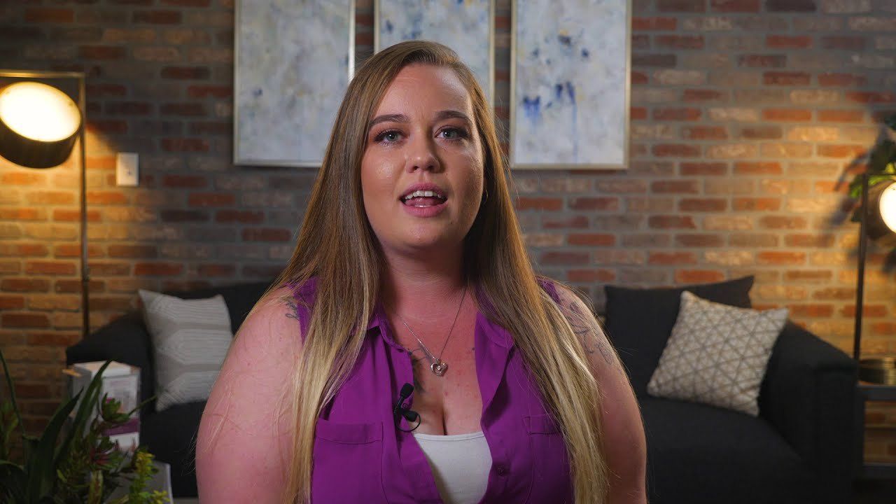 Casey, a Vein Envy patient with long blonde hair and a purple top, talks on a couch in front of a brick wall.