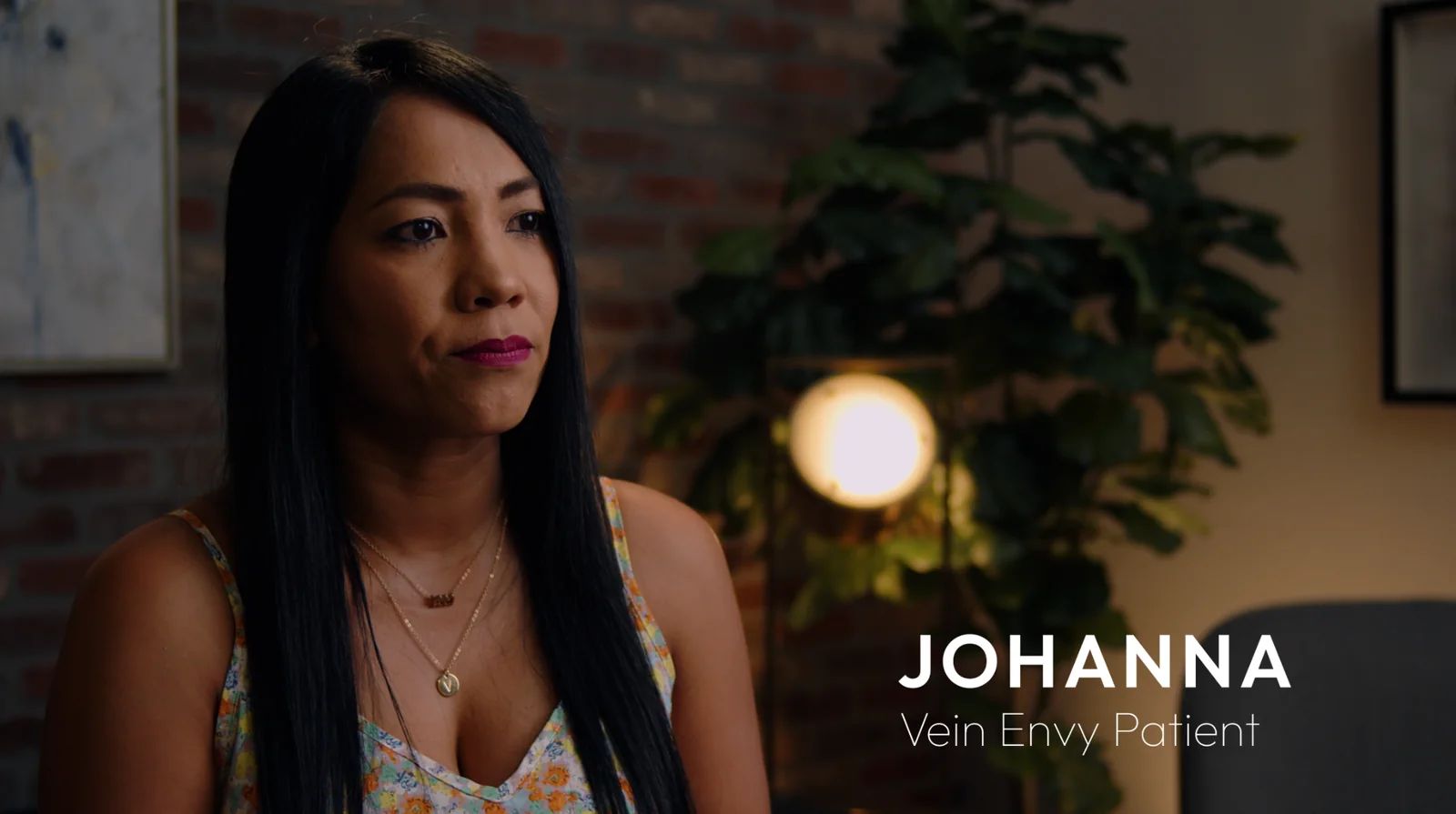 Johanna, a Vein Envy patient with long dark hair, tells her story on camera in front of a brick wall and a lamp.