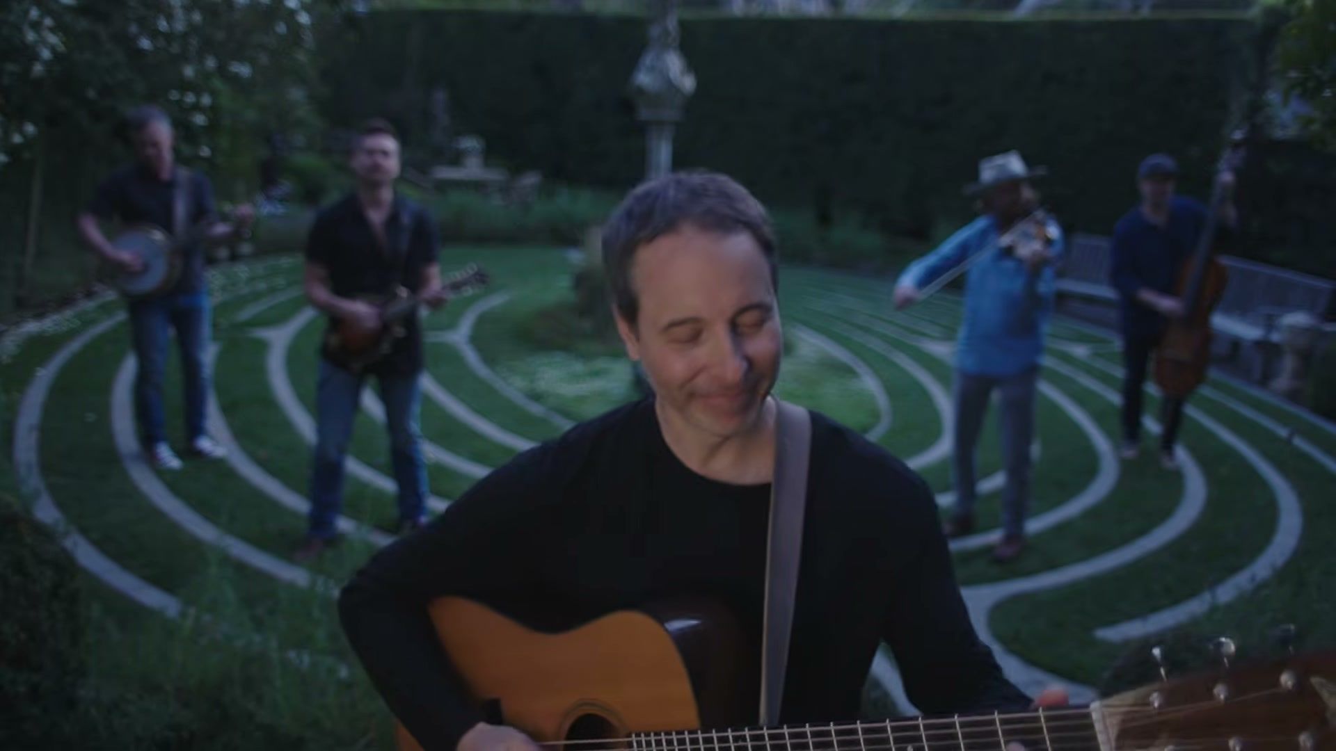 Yonder Mountain String Band play on a garden labyrinth at dusk, a guitarist close in the foreground.