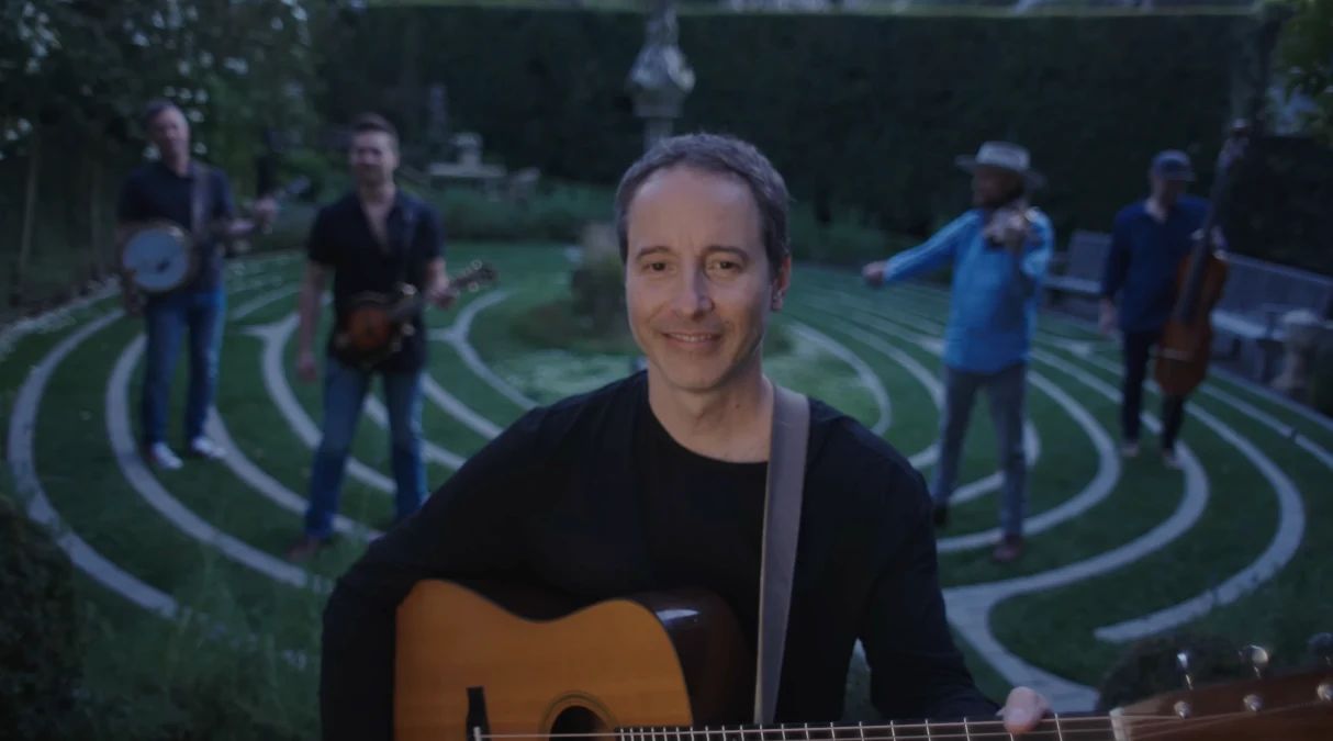 A guitarist sings in a garden labyrinth at dusk, his bandmates playing behind him.